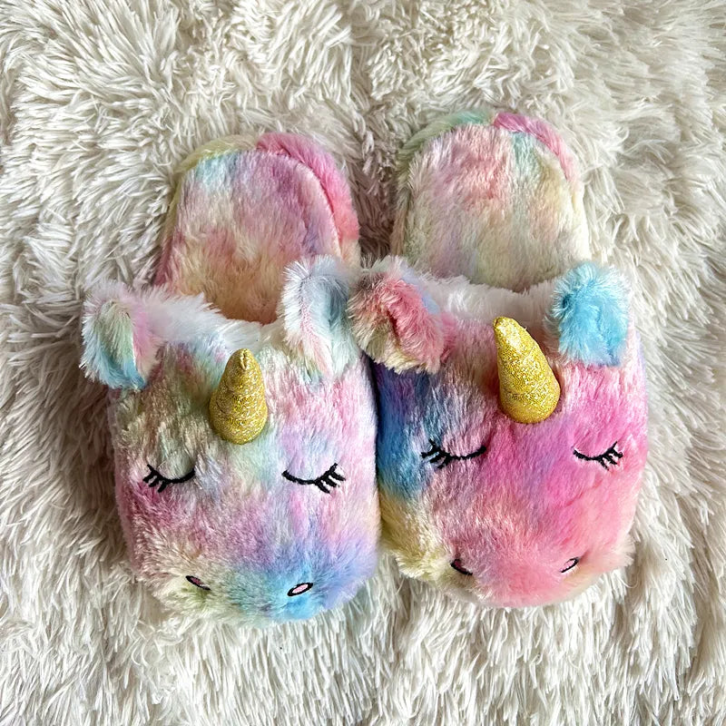 Plush rainbow slip-on slippers shaped like unicorns, featuring their face on the front with closed eyes, embroidered nostrils, and a golden horn.